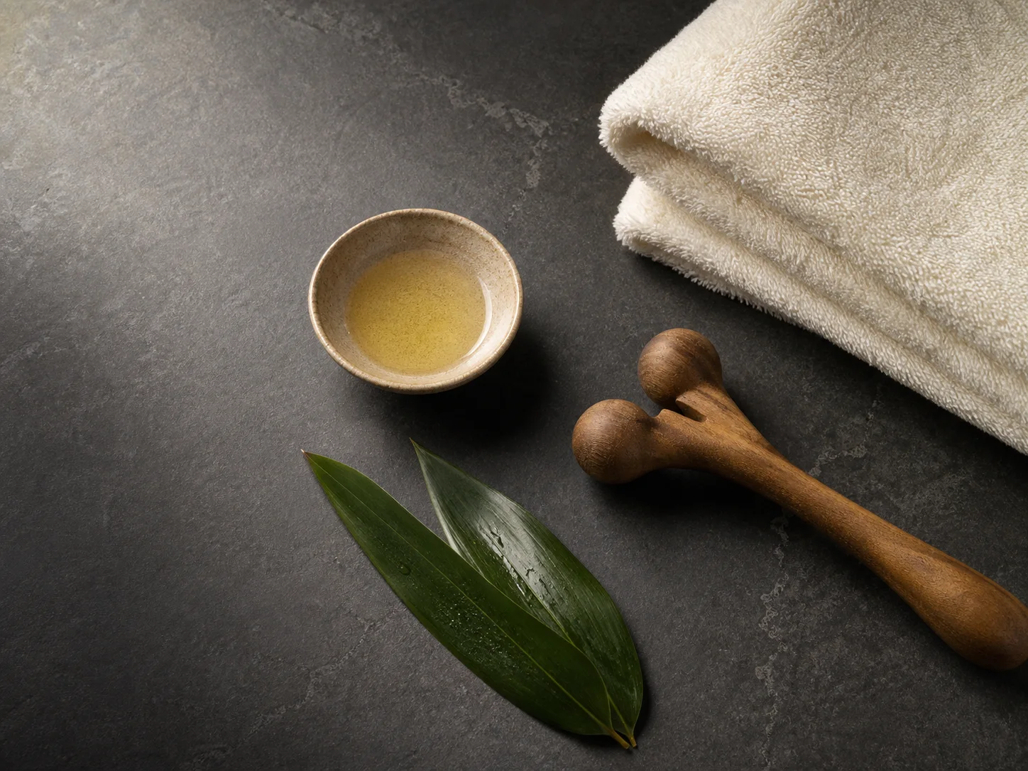 Towel, neutral oil and a wooden massage tool on dark stone
